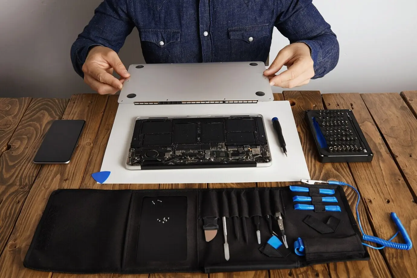 Servicetechniker öffnet die rückseitige Topcase-Abdeckung eines Laptop-Computers, bevor er ihn mit seinem professionellen Werkzeug aus dem Werkzeugkasten in der Nähe auf einem Holztisch repariert, reinigt und instand setzt, in Vorderansicht.